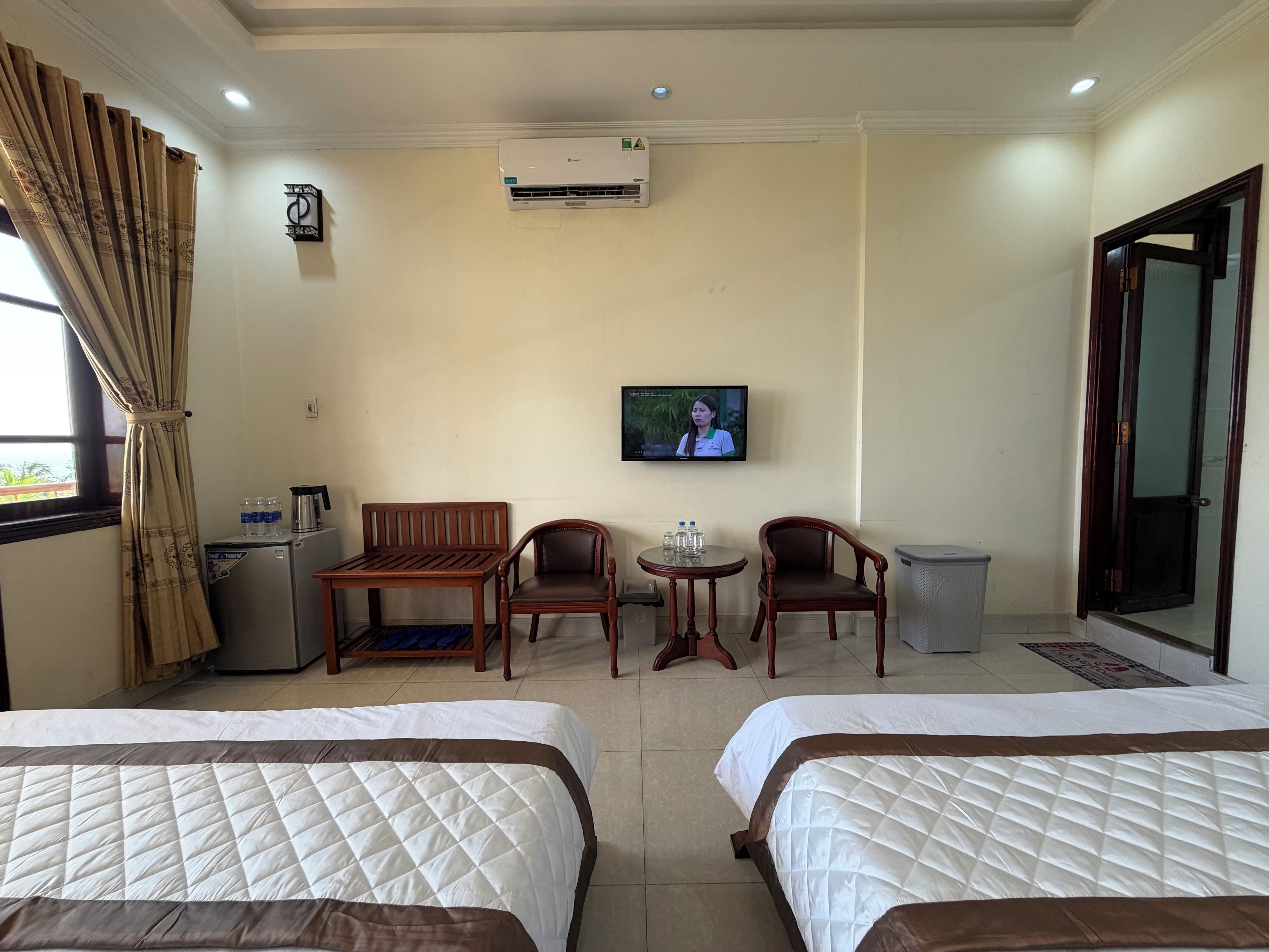 A hotel room with two beds, a TV, a small table with chairs, a refrigerator, and an air conditioning unit.