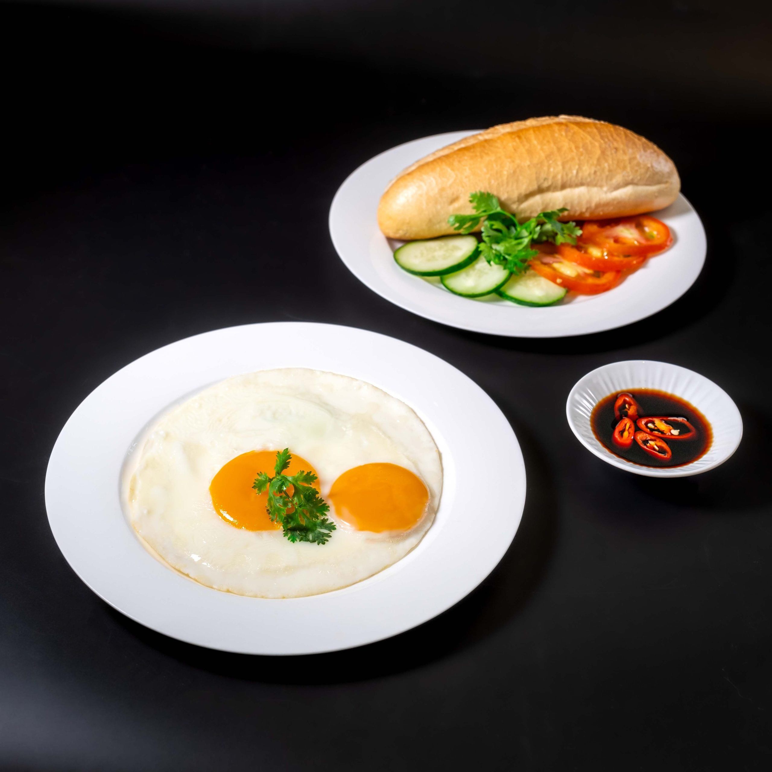 Prato com dois ovos ensolarados, decorado com salsa, um sanduíche com legumes e uma pequena tigela de molho.
