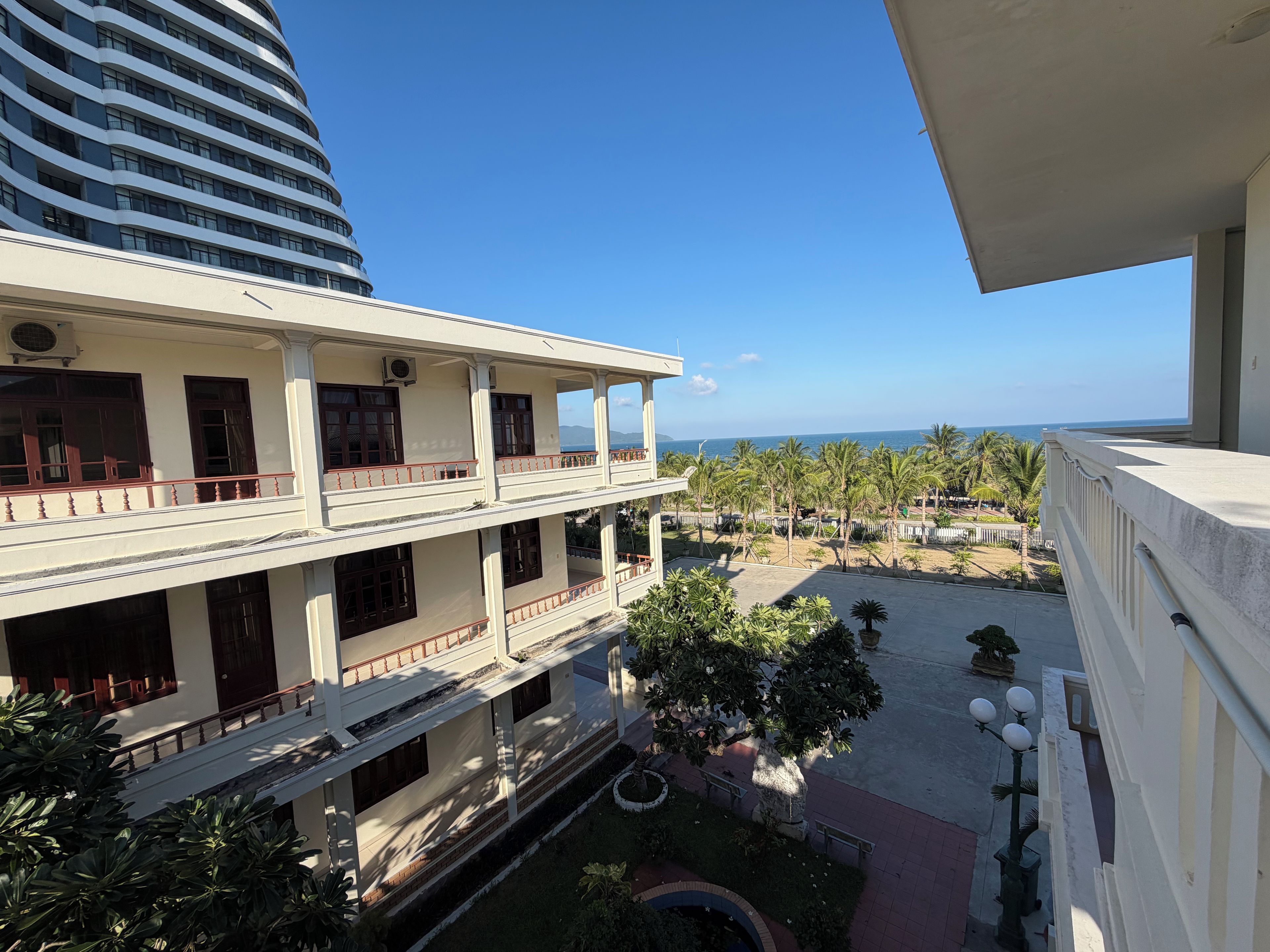 A multi-story building with balconies overlooking a landscaped area with palm trees and a view of the sea.