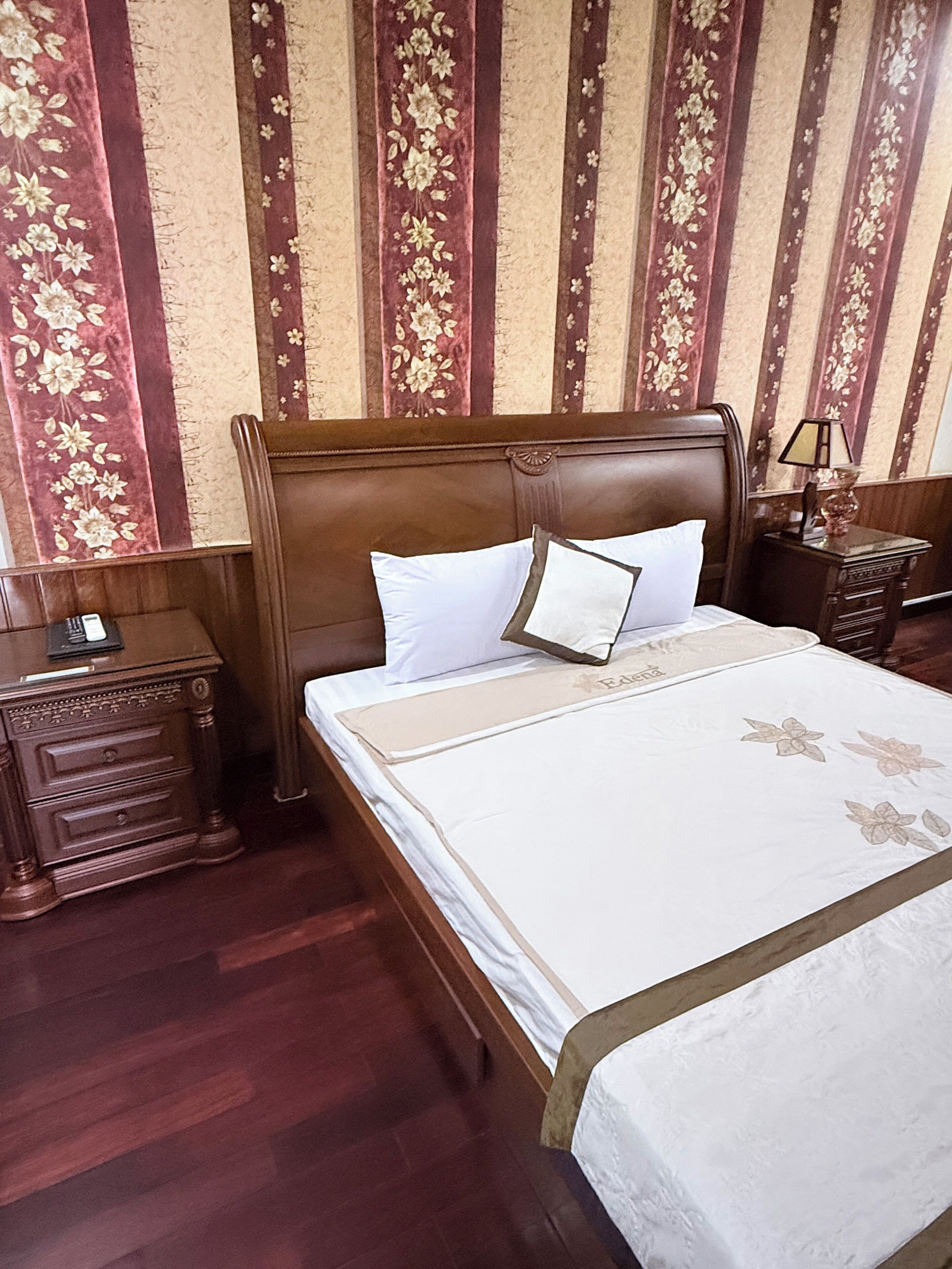 A neatly made bed with a wooden headboard, a bedside table with a lamp, and a floral-patterned wall.