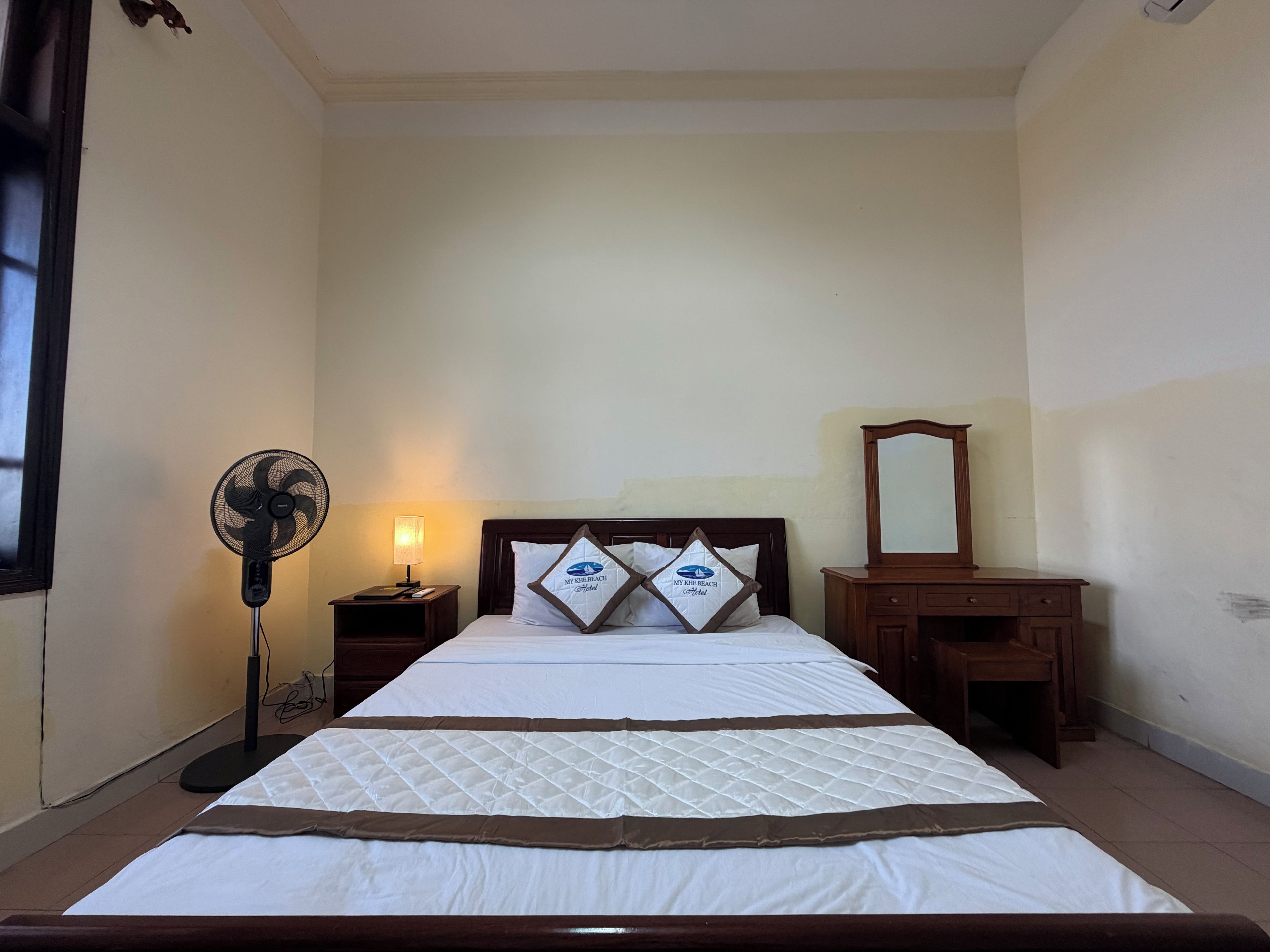 Une chambre d’hôtel avec un lit, une lampe de chevet, un ventilateur sur pied, une table de chevet et une commode en bois.