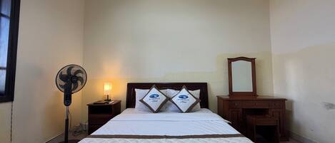 Une chambre d’hôtel avec un lit, une lampe de chevet, un ventilateur sur pied, une table de chevet et une commode en bois.