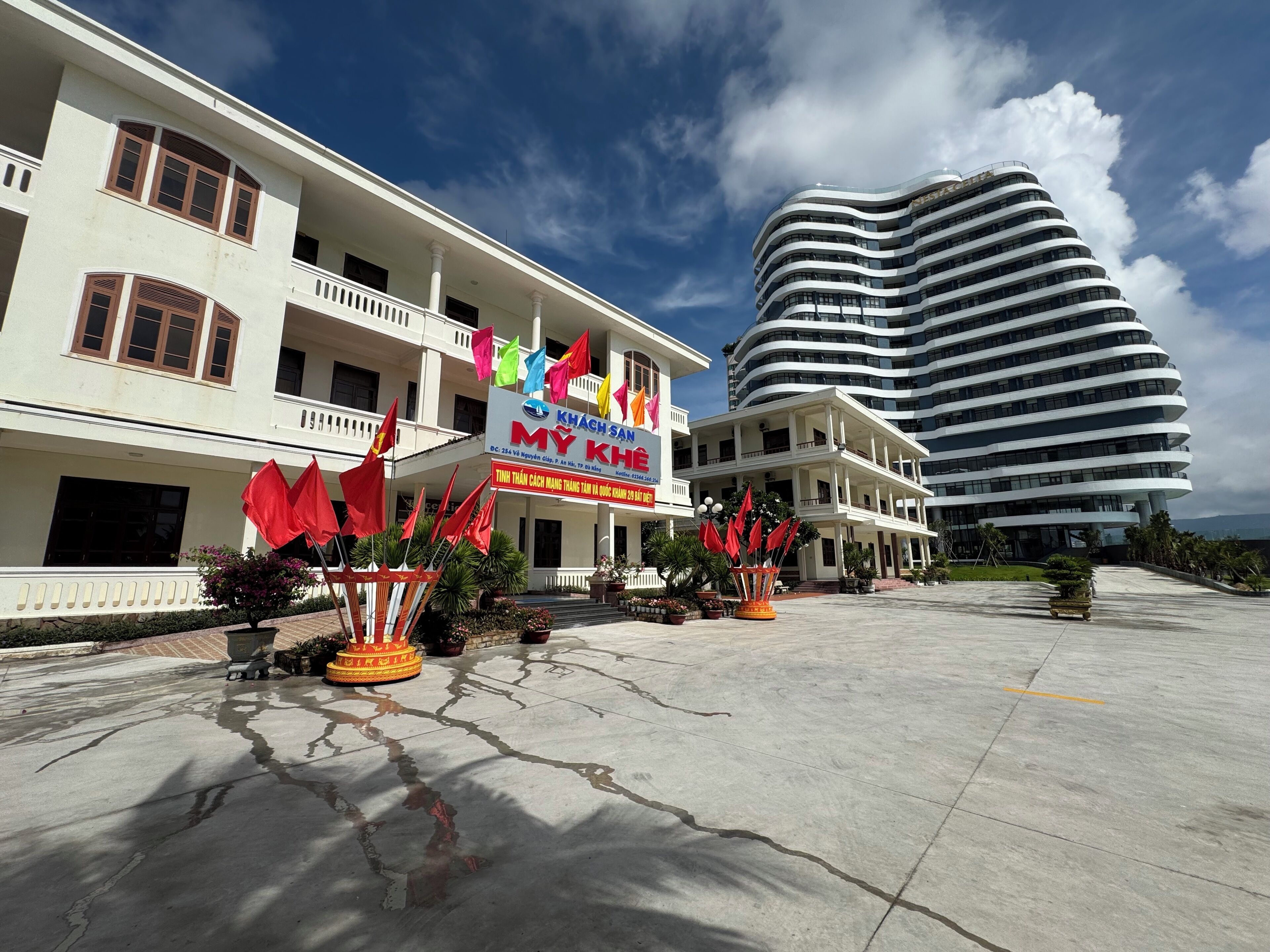 A hotel with a traditional and a modern building. The hotel has a signboard with flags and a large parking area.