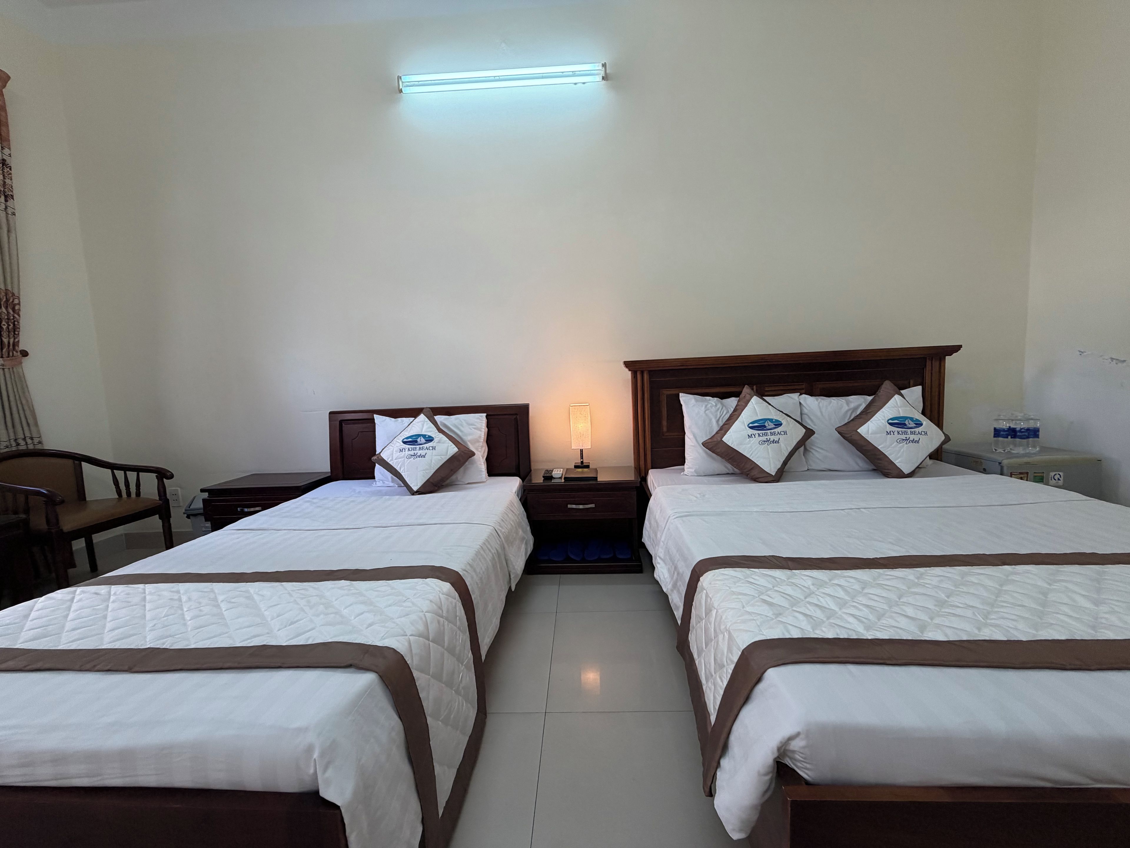 Une chambre d’hôtel avec deux lits, une table de chevet avec une lampe, une chaise et un distributeur d’eau.