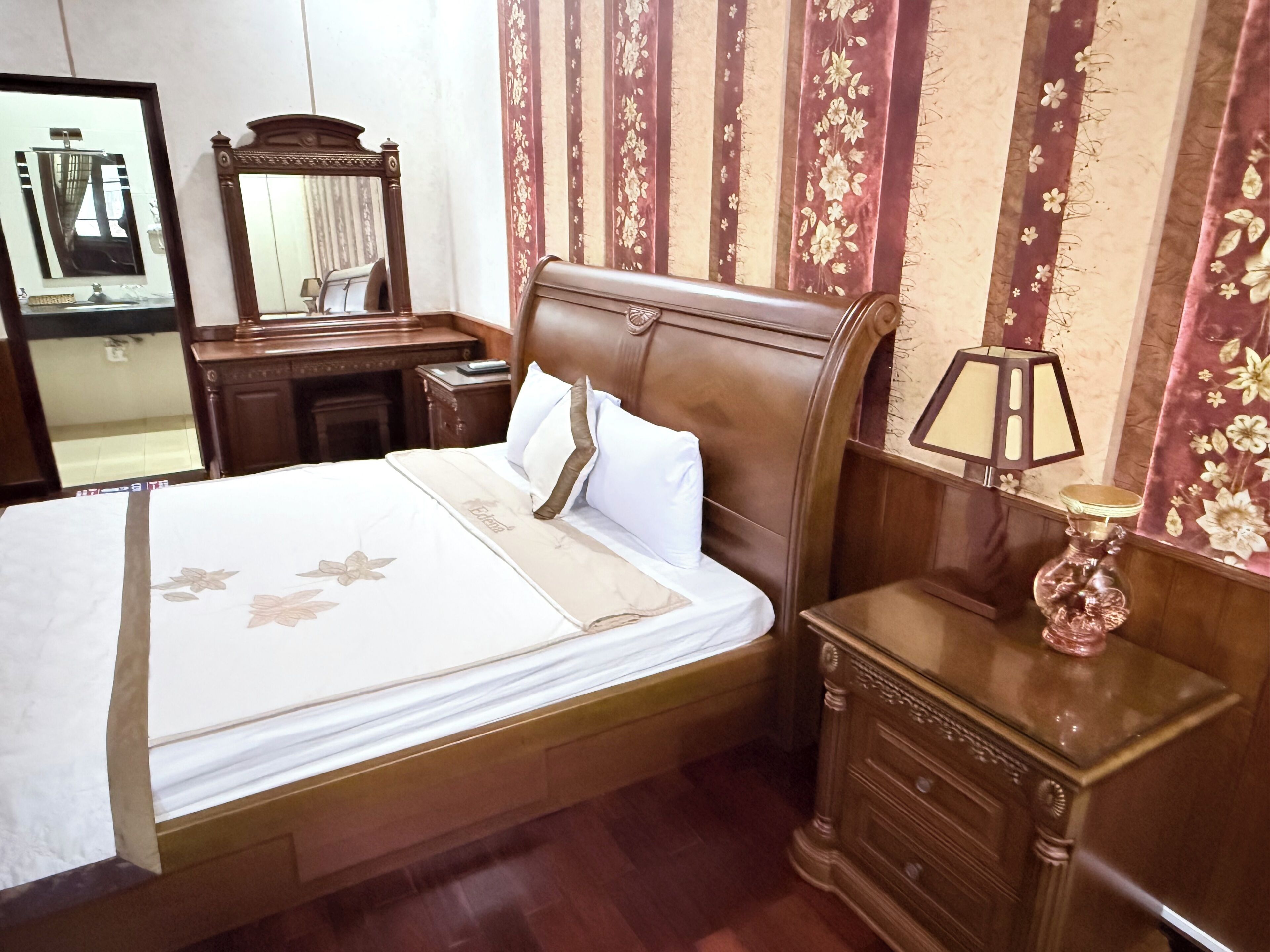 A neatly made bed with a floral bedspread, a wooden nightstand with a lamp, and a mirror on the wall.