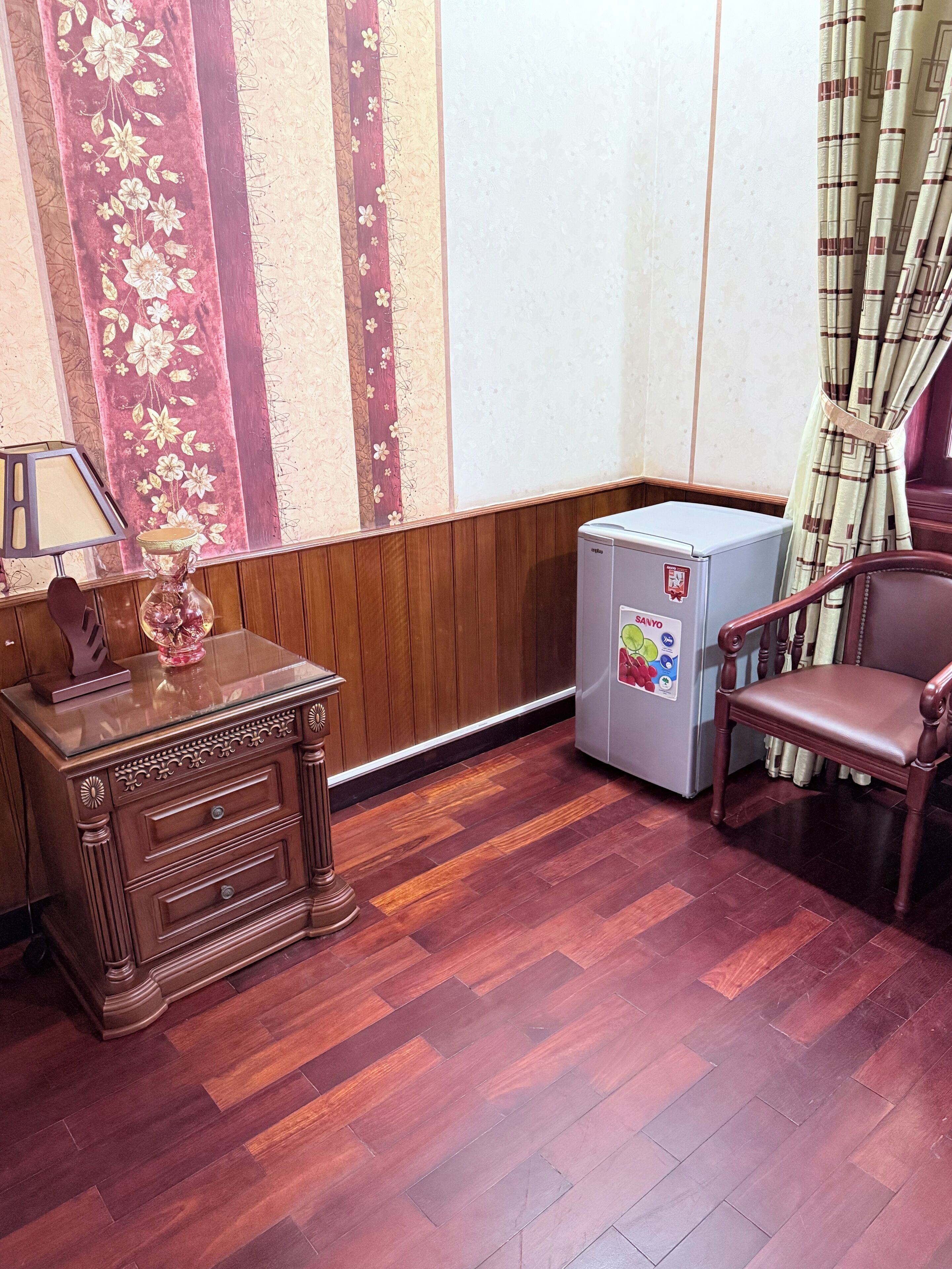 A corner room with a wooden cabinet, a small refrigerator, a chair, and a window with curtains.