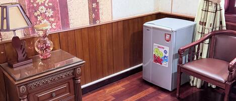 A corner room with a wooden cabinet, a small refrigerator, a chair, and a window with curtains.