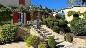 A traditional Mediterranean-style building with a stone staircase leading to a white door, surrounded by well-maintained gardens and a clear blue sky. - Charming family home with heated pool in the Gard (Méjannes Lès Alès)