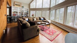 A spacious living room with a large brown sofa, a coffee table, and a red patterned rug. There is a bookshelf filled with books and decorative items, and a dining area with chairs and a table. Large windows provide a view of the outdoors. - Mountain View, Dogs OK, 2 Kitchens, Screened Deck, Pool Table, North Conway (Conway)