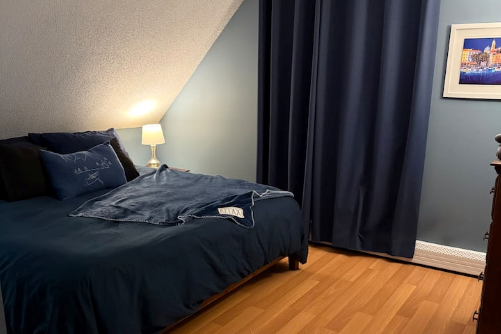 Un dormitorio con cama, una mesita de noche, lámpara, cortina oscura y un cuadro en la pared.