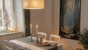 Un comedor con una mesa blanca, cuatro sillas, una lámpara colgante, un cuadro en la pared y una ventana con cortinas.