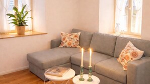 Una sala de estar acogedora con un sofá seccional gris, dos mesitas blancas con candelabros geométricos, una lámpara colgante y una planta en maceta en el alféizar de una ventana.