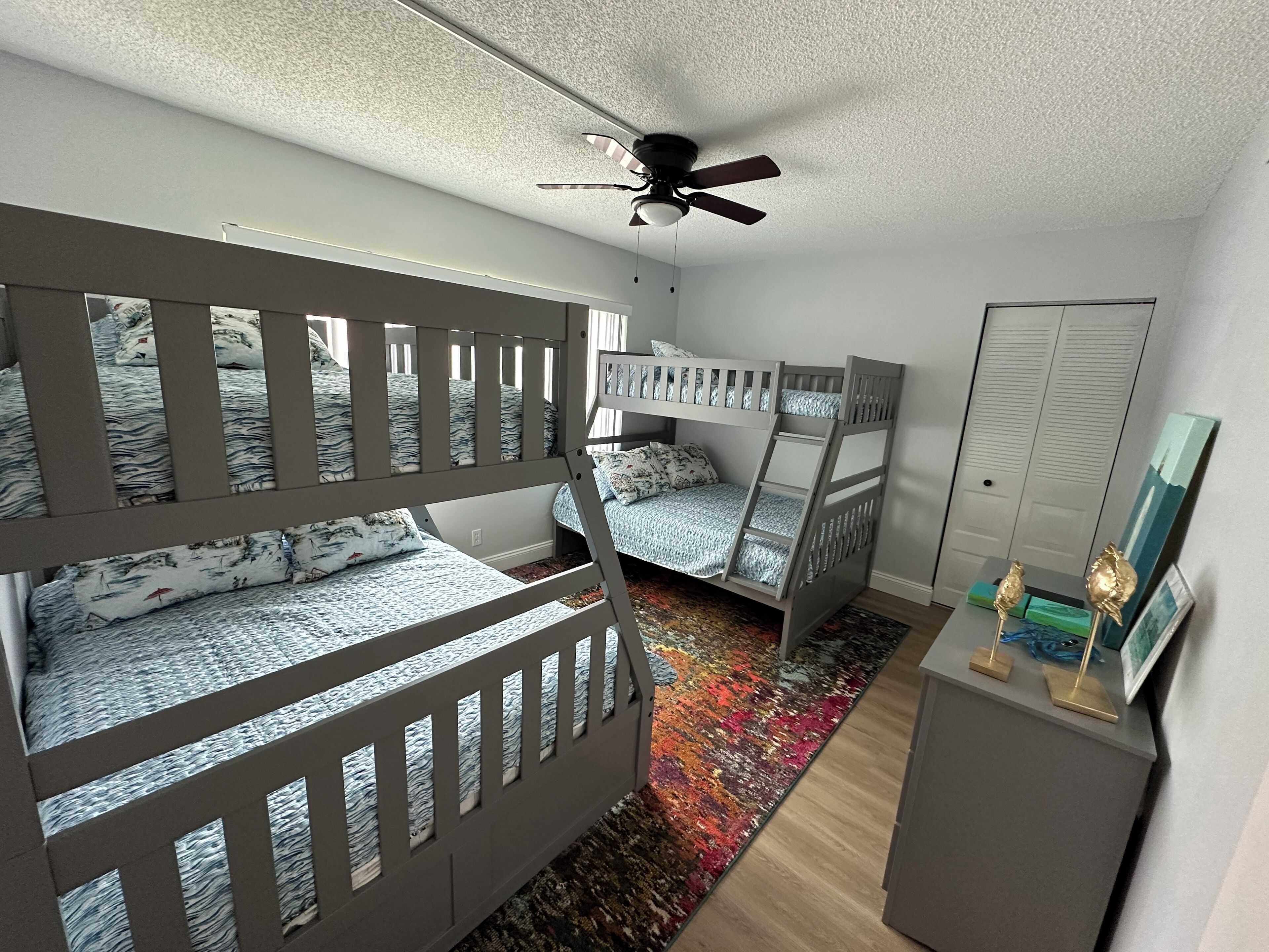 Une chambre avec des lits superposés, un ventilateur de plafond, un tapis coloré et un petit bureau agrémenté d’objets décoratifs.