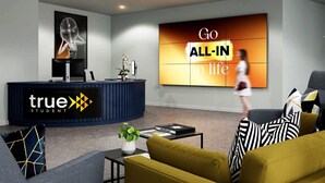 A modern reception area with a curved desk, a large screen displaying "Go ALL-IN on life", and a seating area with sofas and a coffee table. - Student Only True Leicester (Leicester)