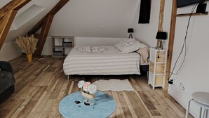 Un dormitorio acogedor en el ático, con techo inclinado, cama con ropa de cama a rayas, una mesita de noche blanca pequeña, un televisor montado en la pared y una mesa redonda azul con un jarrón de flores.
