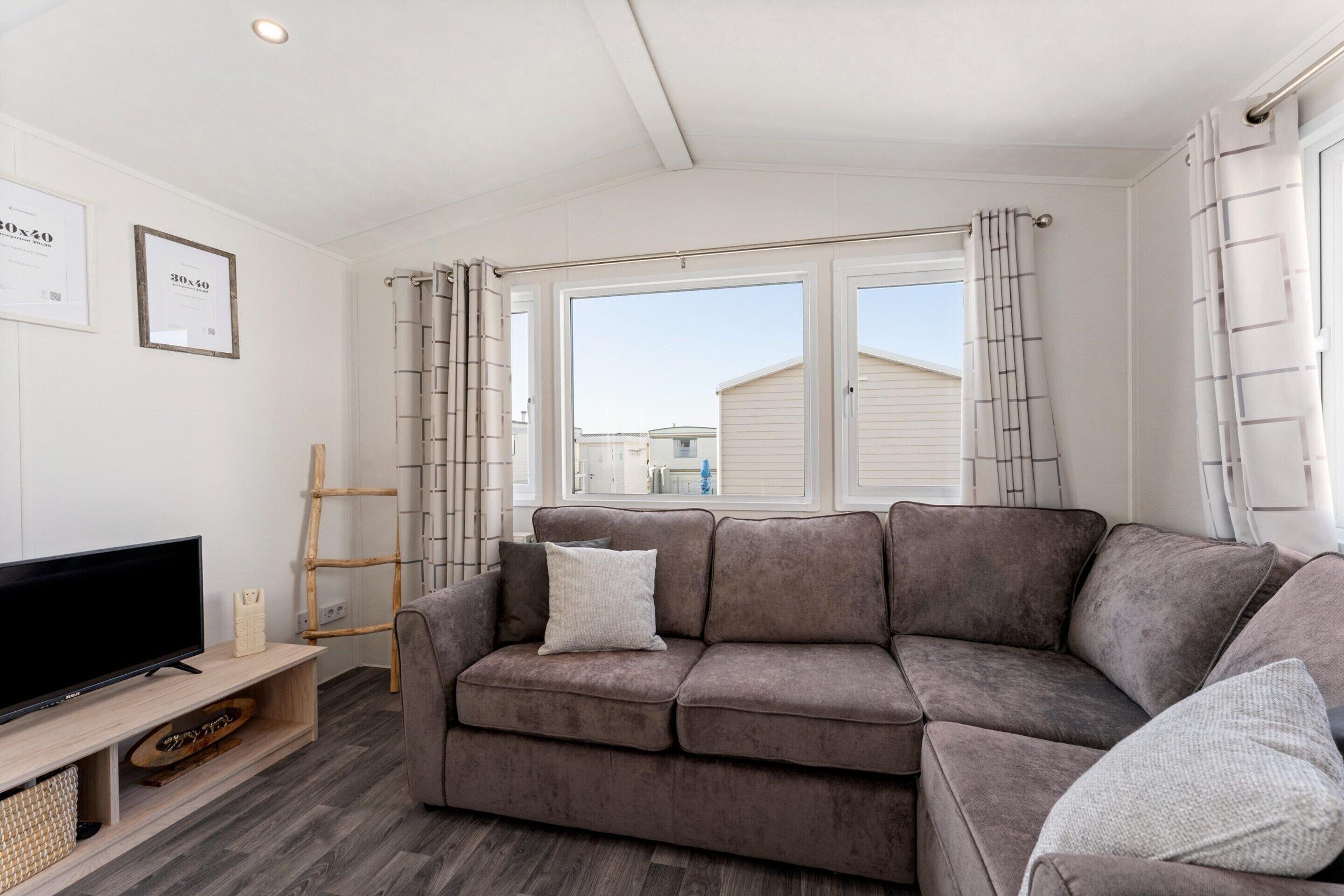 A living room with a grey sofa, a TV on a wooden stand, a ladder, and a window with curtains.