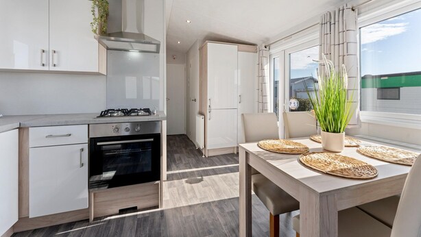 A modern kitchen with white cabinets, a gas stove, and an oven. There is a dining table with woven placemats and a potted plant. - Comfortable Mobile Home in Middelkerke (Middelkerke)