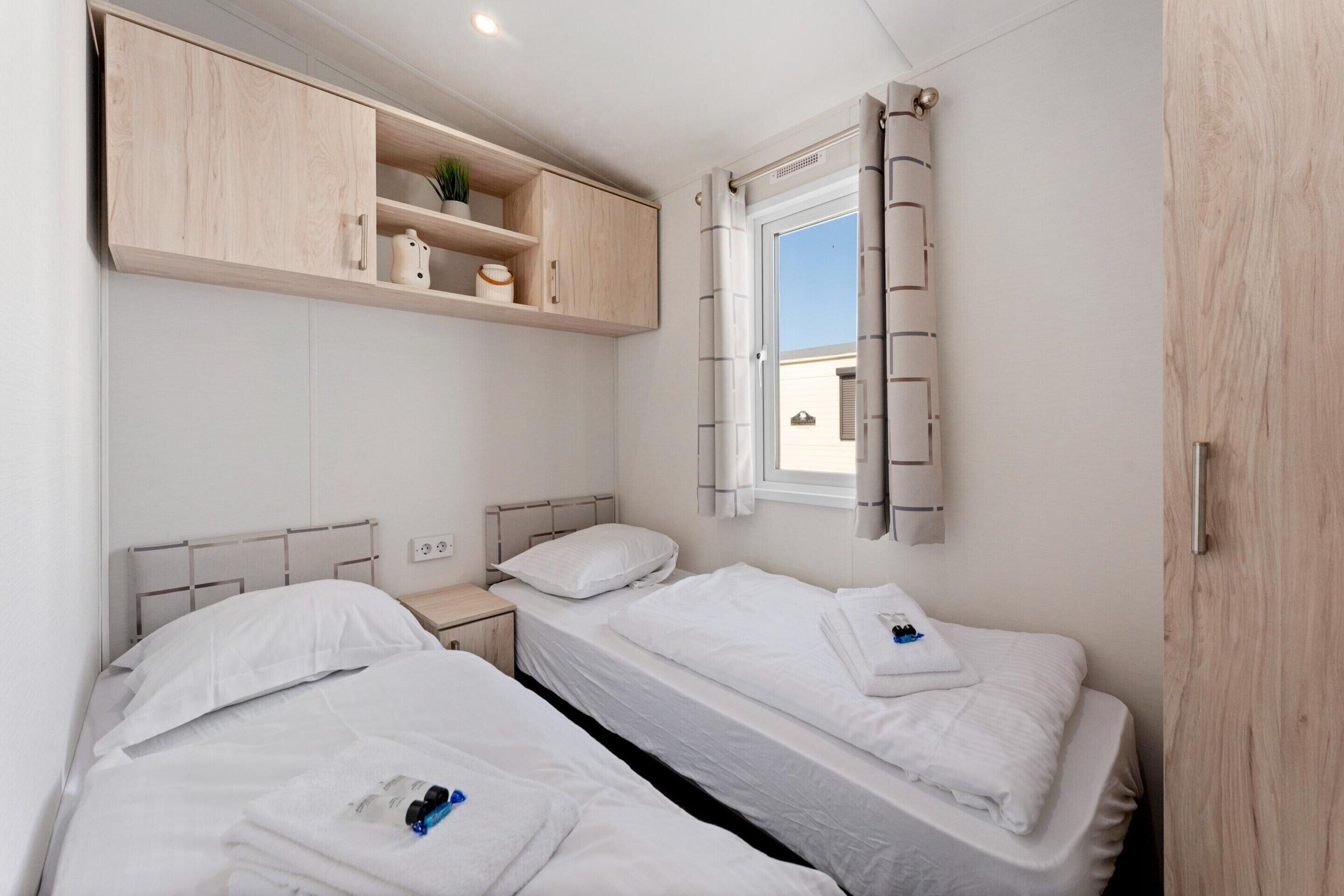 Une chambre d’hôtel compacte, dotée de deux lits simples, d’une armoire en bois, d’une petite table de chevet et d’une fenêtre avec des rideaux.