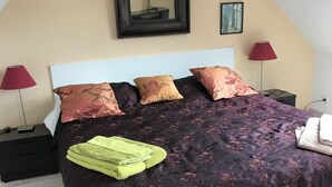 Ein ordentlich bezogenes Bett mit einem dunkelblumenmuster-Bettüberwurf, mehreren Kissen und einem grünen Handtuch. Zwei Nachttische mit Lampen, ein gerahmter Spiegel und ein kleines Bild an der Wand sind ebenfalls zu sehen.