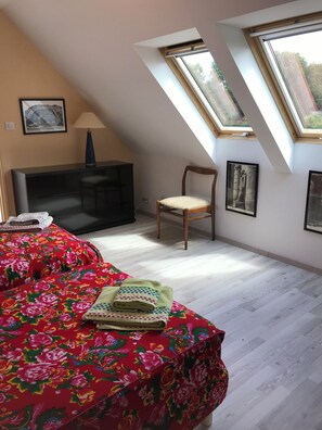 Un dormitorio con techo inclinado, una cama con cubrecama floral, una silla, un escritorio con lámpara y dos ventanas.