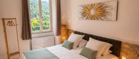 A bedroom with a bed, a hanging light fixture, a window with curtains, a wall-mounted artwork, and a bedside table with a lamp.