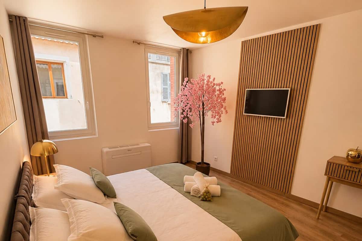 A bedroom with a bed, a bedside table, a lamp, a window, a wall-mounted TV, and a potted plant.