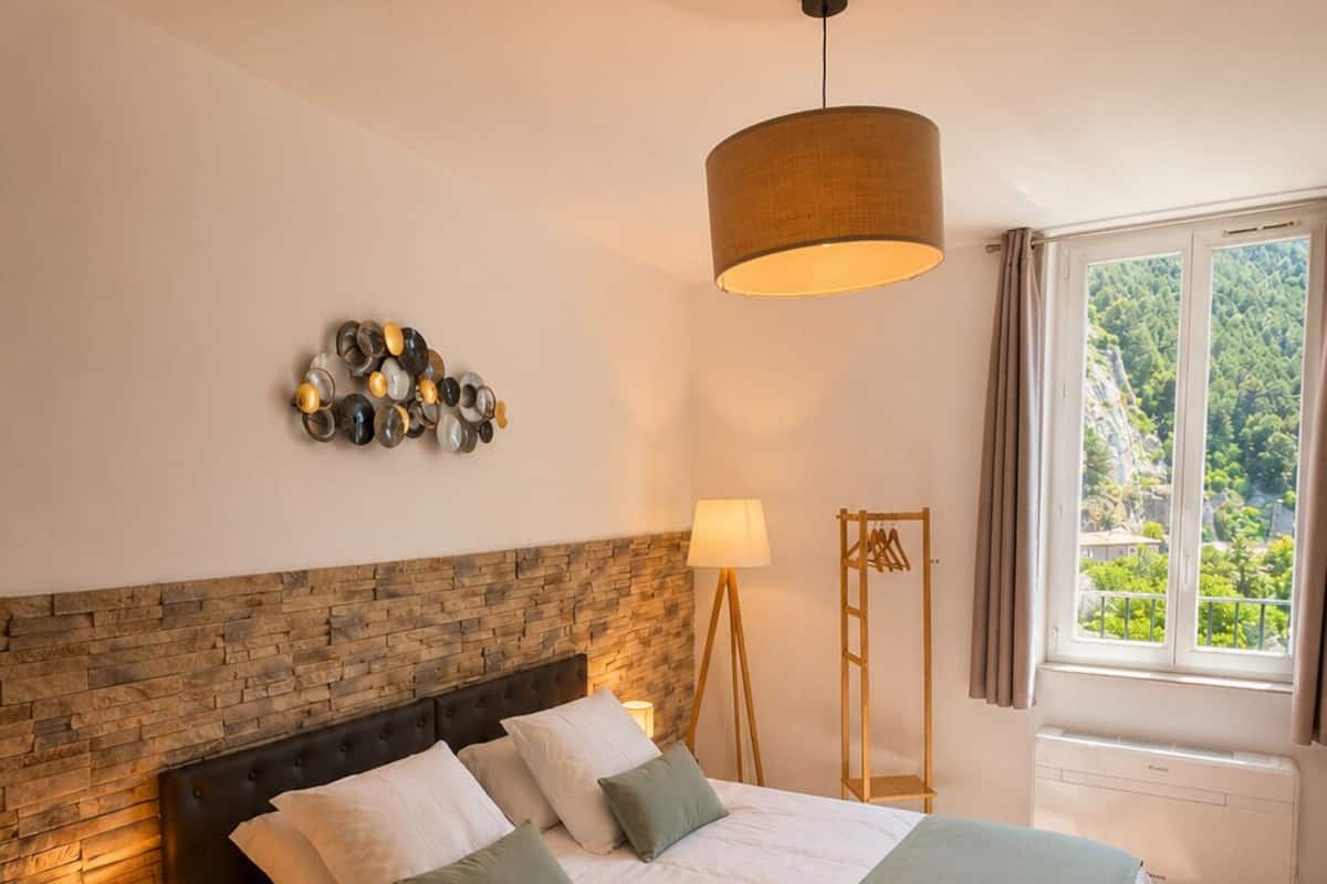 A bedroom with a bed, a nightstand, a lamp, a wall-mounted light fixture, a window with a view, and a decorative wall piece.