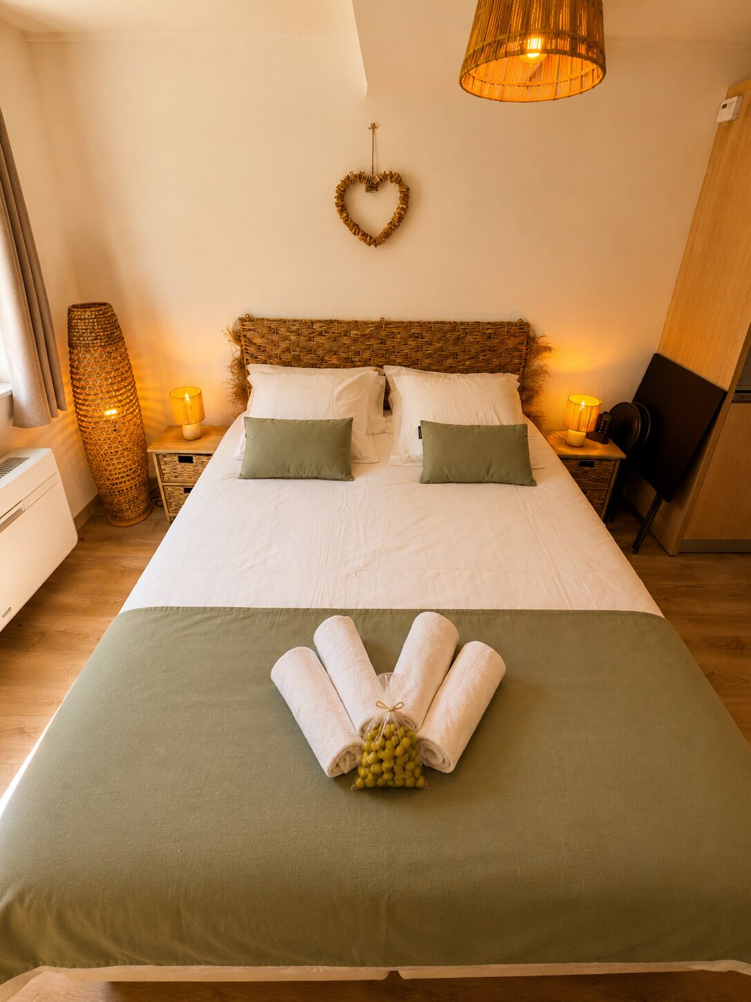 Un lit bien fait, agrémenté d’une décoration en forme de cœur, d’un plaid vert et d’une serviette roulée, avec une petite botte d’olives posée sur le lit.