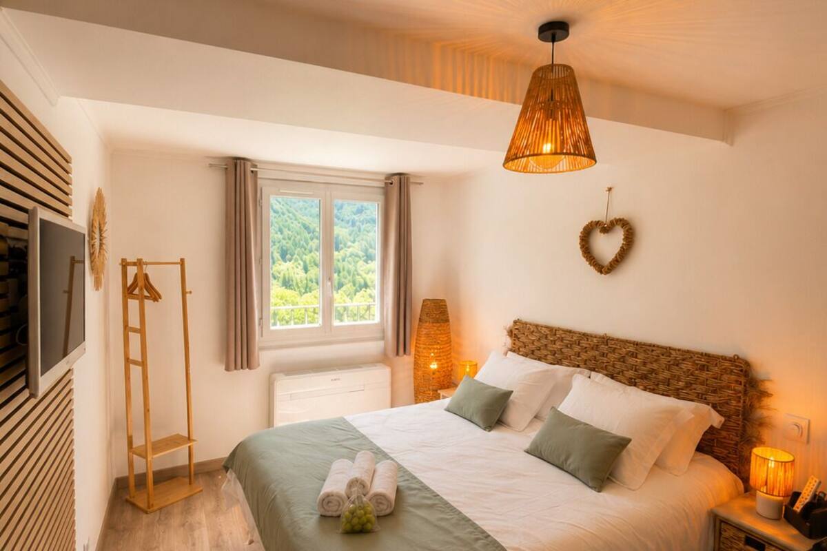 A bedroom with a bed, a bedside table, a lamp, a hanging light fixture, a window, and a wall-mounted TV.
