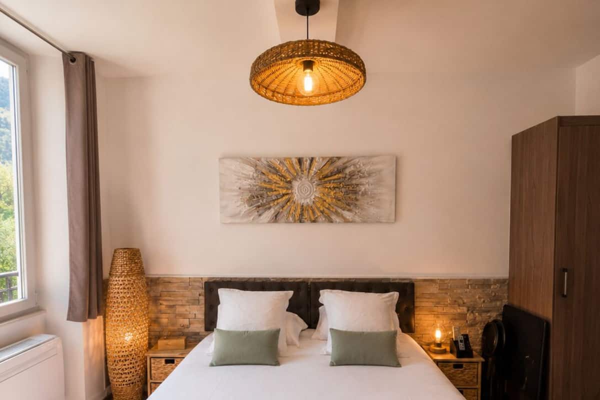A hotel room with a bed, a large woven light fixture, a wall-mounted artwork, and a wooden wardrobe.