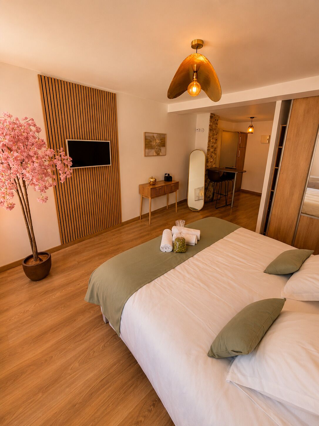 A hotel room with a bed, a wooden wall panel, a TV, a mirror, a small table, and a potted plant.