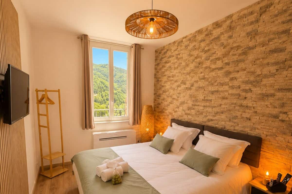 A bedroom with a bed, a TV, a window with curtains, a wooden floor, and a brick wall.