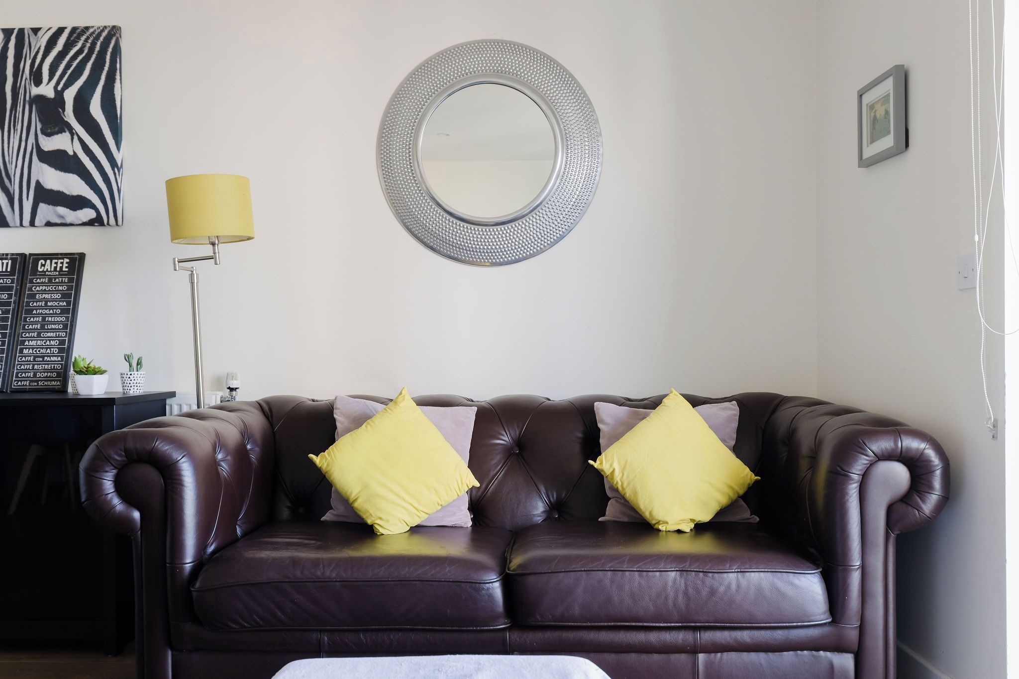 En mørkebrun lædersofa med to gule puder, et rundt spejl, et kunstværk med zebra-mønster, en gul lampe og en menutavle.
