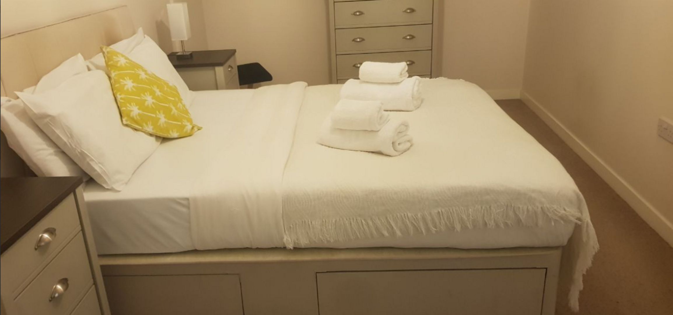 A neatly made bed with white bedding and a yellow pillow, a bedside table with a lamp, and a dresser with drawers.