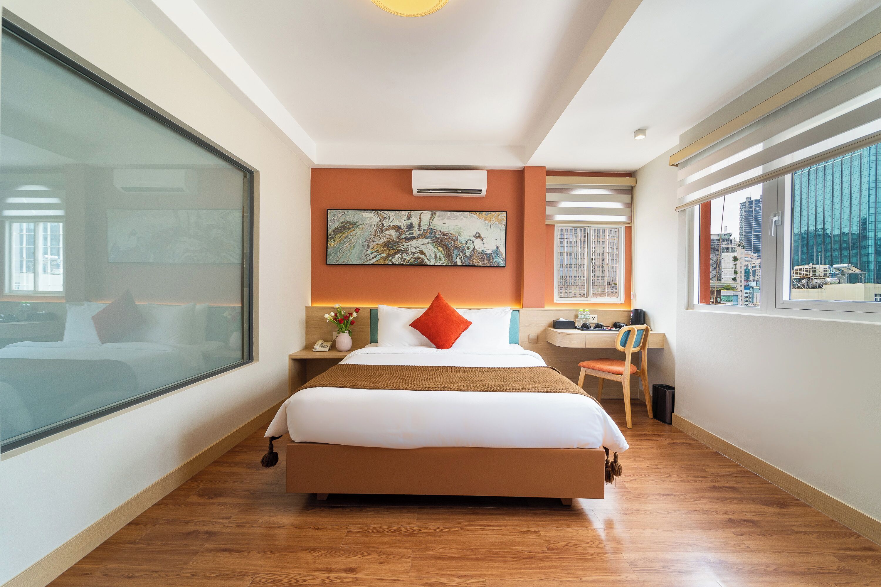 A hotel room with a large bed, a desk, a chair, a window with a city view, and an air conditioning unit.