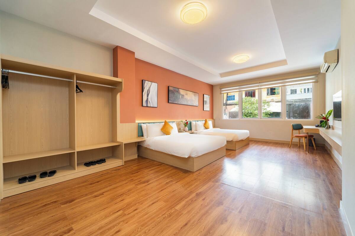 A modern hotel room with a wooden floor, a bed with white bedding, a desk with a chair, and a window with a view of buildings.