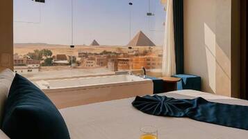 Una habitación de hotel con un ventanal grande, una cama, una bandeja con desayuno y vistas a las pirámides.