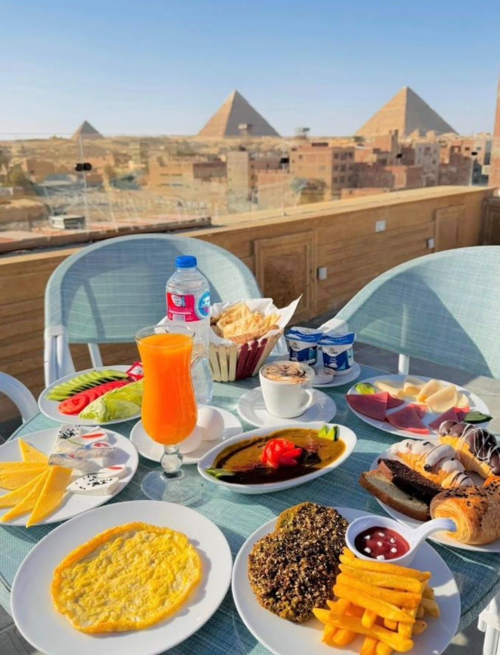 Una mesa dispuesta con diversos alimentos para el desayuno, como huevos, pan y fruta, con vistas a las pirámides al fondo.