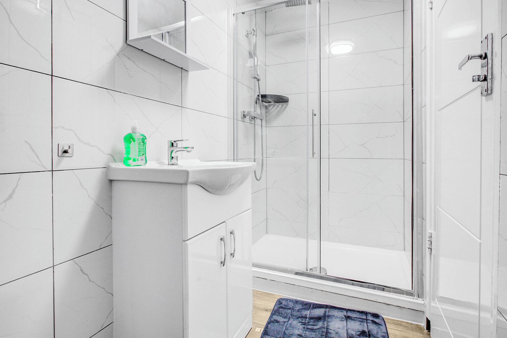 Un baño moderno con baldosas de mármol blanco, cabina de ducha de vidrio, un lavabo con una botella verde y una alfombra azul en el suelo.