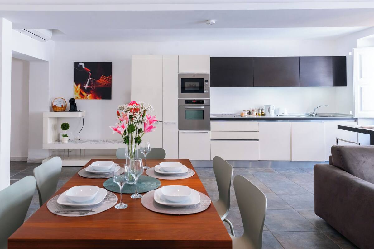 A modern dining area with a wooden table set for four, a vase with flowers, and a kitchen in the background.