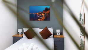 A neatly made bed with white linens and brown pillows, a bedside table with a lamp, and a framed picture of a coastal scene on the wall.