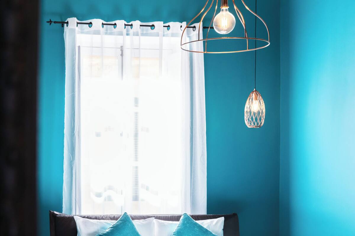 A modern bedroom with a bed, a small bedside table, a window with curtains, and a hanging light fixture.