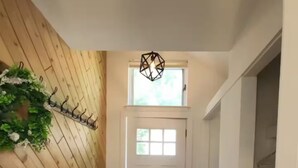 Un couloir avec un banc en bois, un tapis noir, une porte blanche et un luminaire suspendu.
