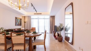 A modern dining area with a round wooden table, cushioned chairs, and a chandelier. There is a large mirror on the wall and a potted plant near the window. - Designer 2BR Serviced Apart at Cairo Festival City (Cairo Governorate)