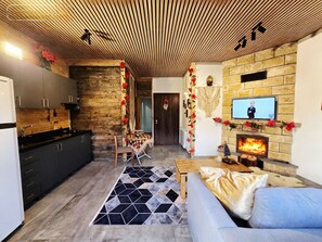 A modern kitchen with wooden accents, a dining area with a patterned rug, a sofa, and a fireplace with a TV above it.