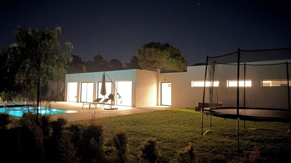 A modern house with a swimming pool, a trampoline, and a well-lit patio area.