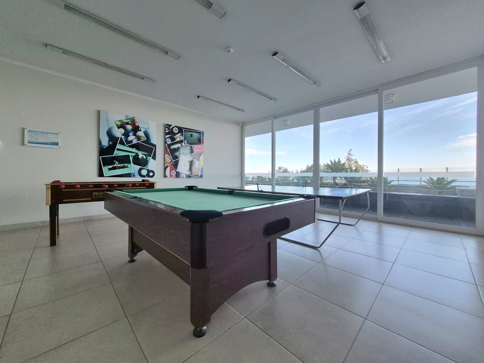 A pool table with a billiards cue, a foosball table, and a large window with a view of the outdoors.