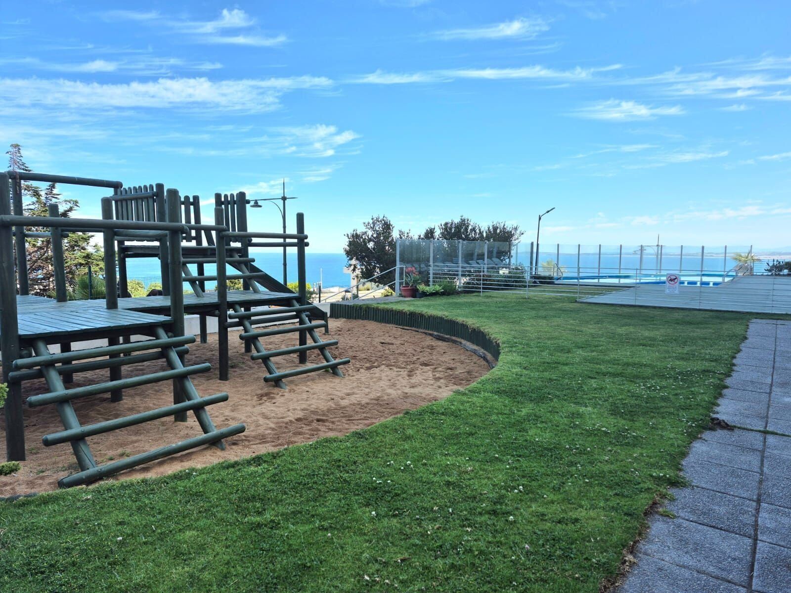 Un parque infantil con estructuras para escalar y una zona de juegos con arena, rodeado de césped y un camino pavimentado.