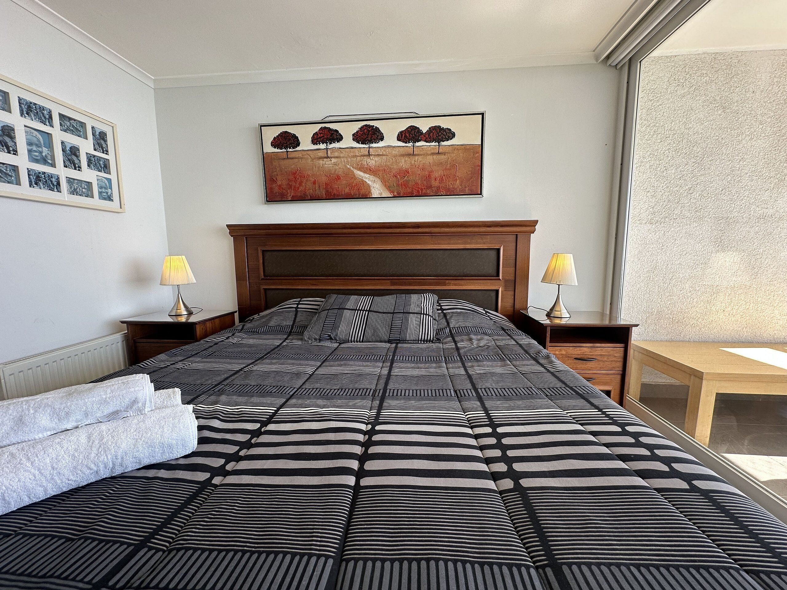 Un lit bien fait, recouvert d’une couette à rayures, avec deux lampes de chevet, un tableau encadré au-dessus du lit et une fenêtre sur le côté droit.
