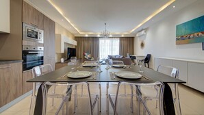 A modern dining area with a long table, clear chairs, and a kitchen in the background. - Swieqi, Luxury Apartmen/3BD- 2BH,sleeps 6 (Is-Swieqi)
